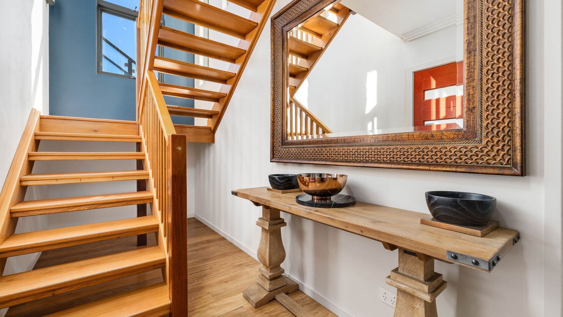 A wooden staircase with a mirror above it, a wooden console table with bowls, and a red door in the reflection.