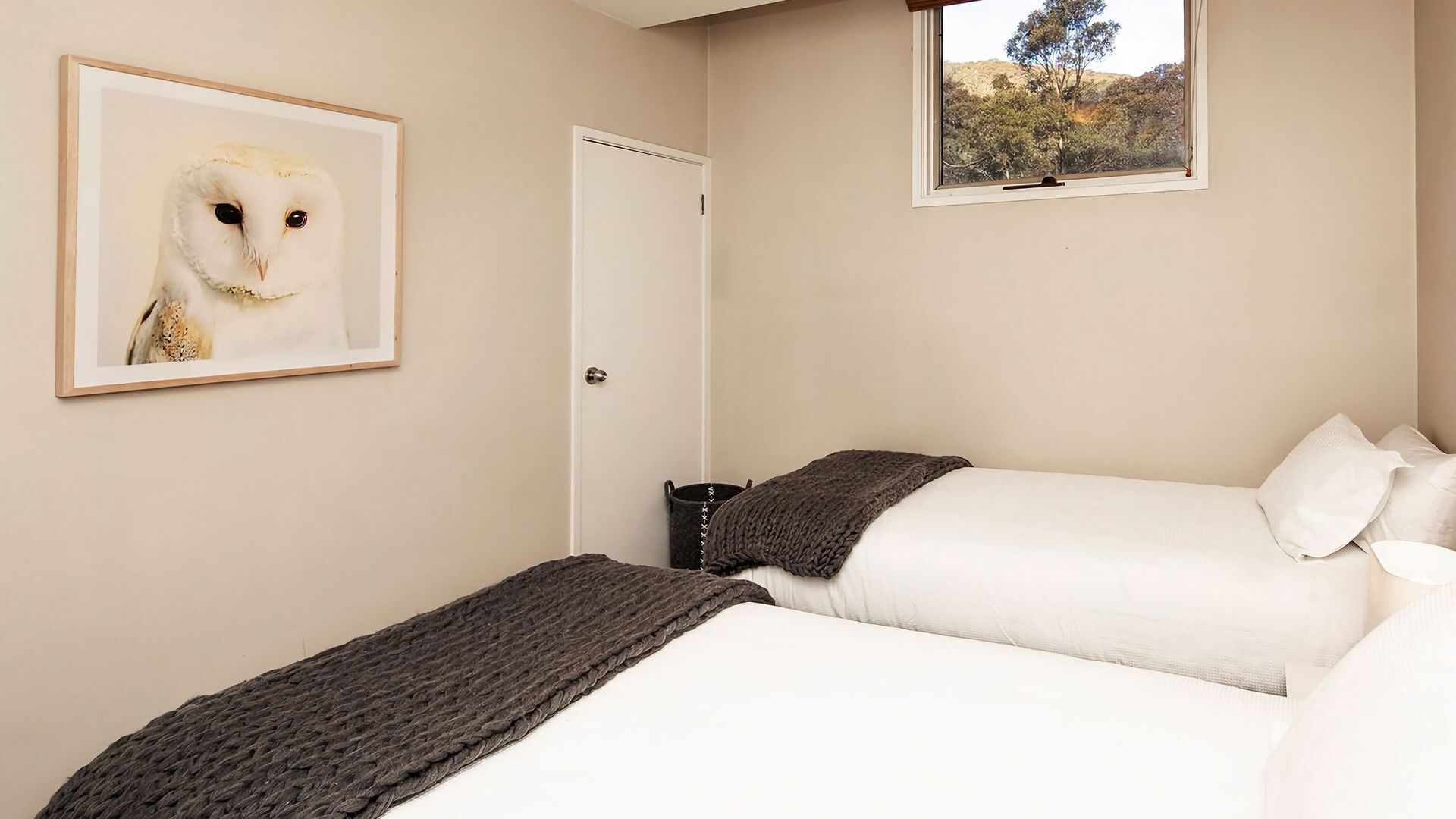 A bedroom with two beds, a framed owl painting, a window with a view of trees, and a door.