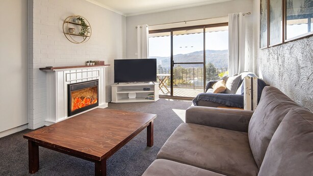 A living room with a fireplace, a flat-screen TV, a sofa, and a balcony with a view of the mountains.