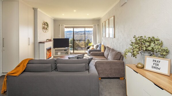 Een moderne woonkamer met een grijze hoekbank, een kleine televisie en een open haard. Er is een balkon met uitzicht op de bergen.