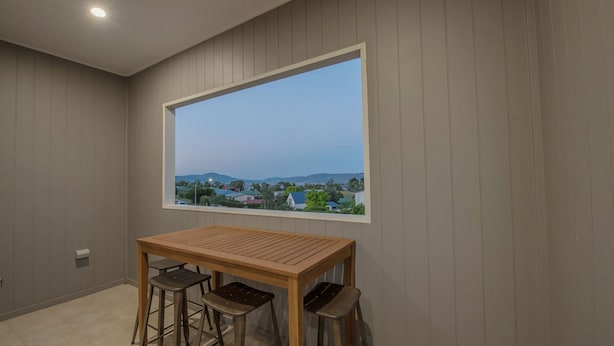 A room with a wooden table and two stools, a window with a view of houses and hills, and a recessed ceiling light.