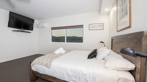 A bedroom with a bed, a TV mounted on the wall, a window with a cityscape view, and a framed picture on the wall.