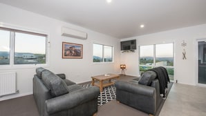 A modern living room with two leather sofas, a wooden coffee table, and a TV mounted on the wall. There are large windows offering views of the countryside.