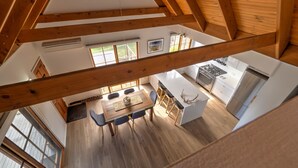 A high-ceilinged dining area with a wooden table and chairs, a kitchen with modern appliances, and large windows allowing natural light.