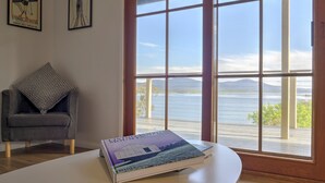 A room with a view of a lake, a grey armchair with a patterned pillow, and a book titled "Living in the Mountains" on a white table.