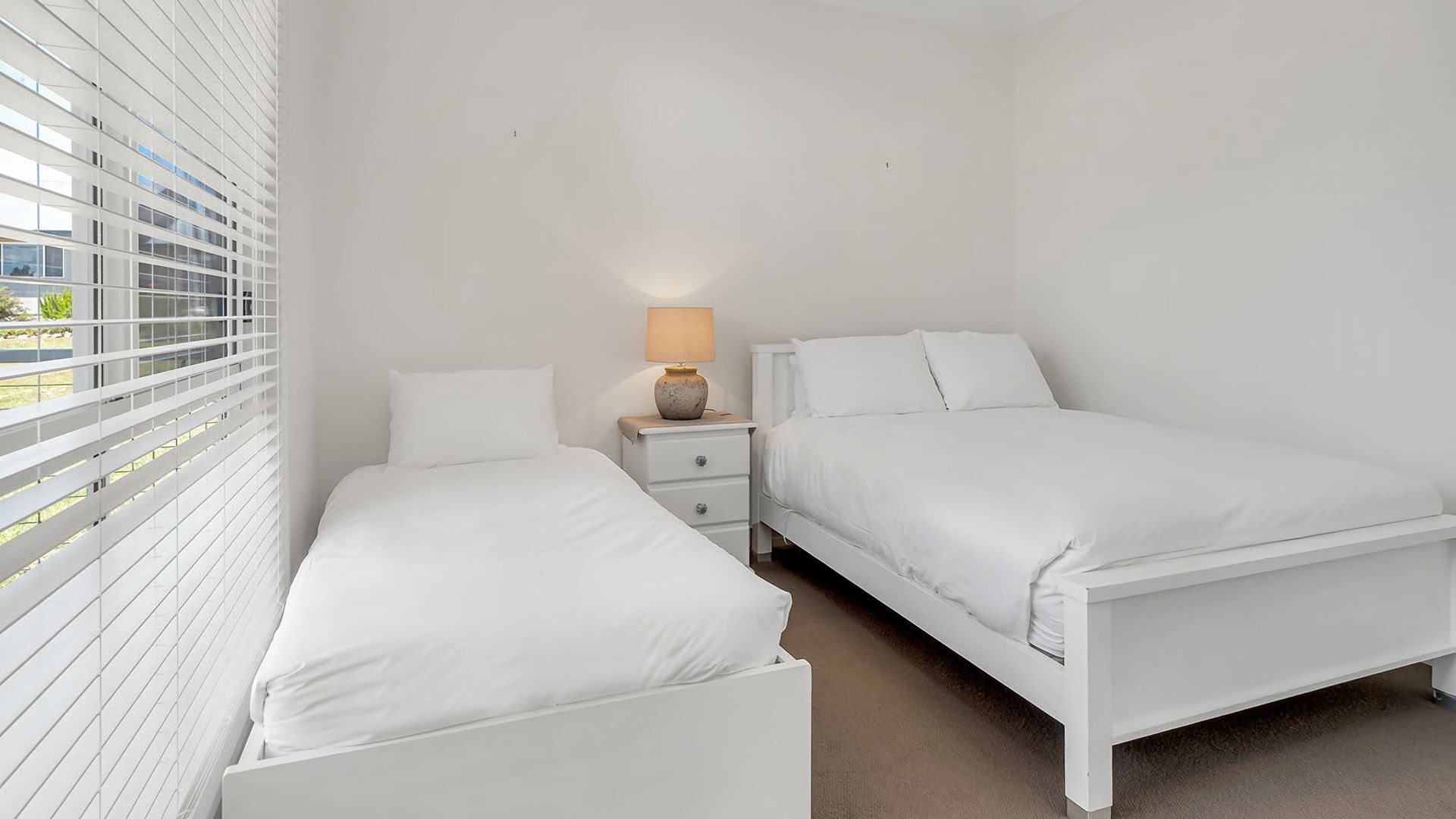 A bedroom with two single beds, a nightstand with a lamp, and a window with blinds.