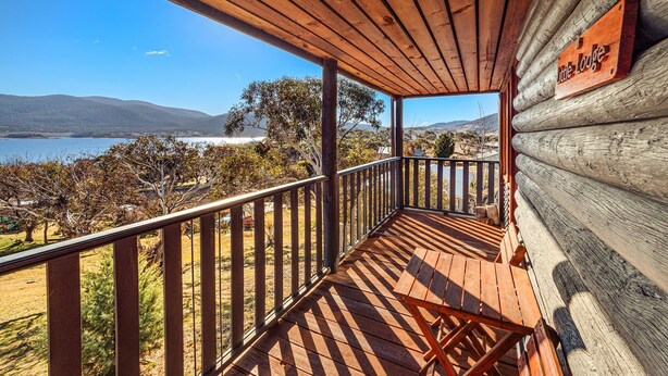Una terraza de madera con barandilla, un letrero que dice "Little Lodge" y vistas a un lago y colinas.