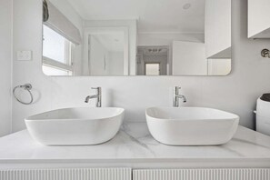 Une salle de bain avec deux vasques blanches, un grand miroir et une porte donnant sur une autre pièce.