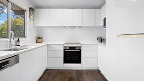 Une cuisine moderne avec des meubles blancs, un four encastré, un évier en acier inoxydable et une fenêtre donnant sur les arbres.