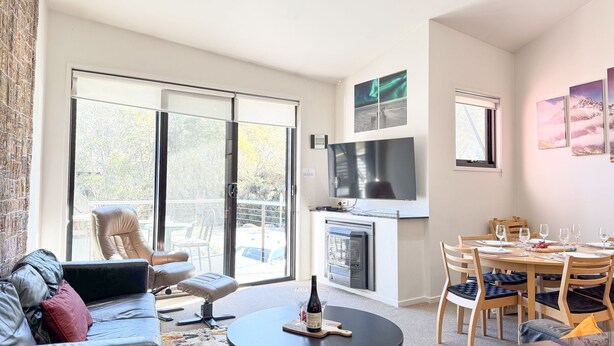 A modern living room with a sofa, a dining table set for four, a fireplace, and a large TV. - Angala 2 located across from Friday Flat (Thredbo)