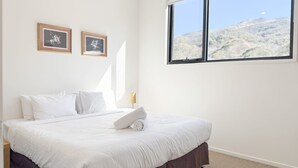 A hotel room with a large bed, two framed pictures on the wall, a window with a mountain view, and a nightstand with a lamp. - Angala 2 located across from Friday Flat (Thredbo)
