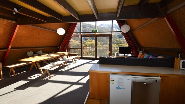 A cozy, well-lit room with a large window offering a scenic view, a wooden table, benches, and a sofa with cushions.