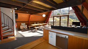 A compact kitchen with a dishwasher, wooden dining table, and chairs, a sofa, and a large window with a view of a mountainous landscape.