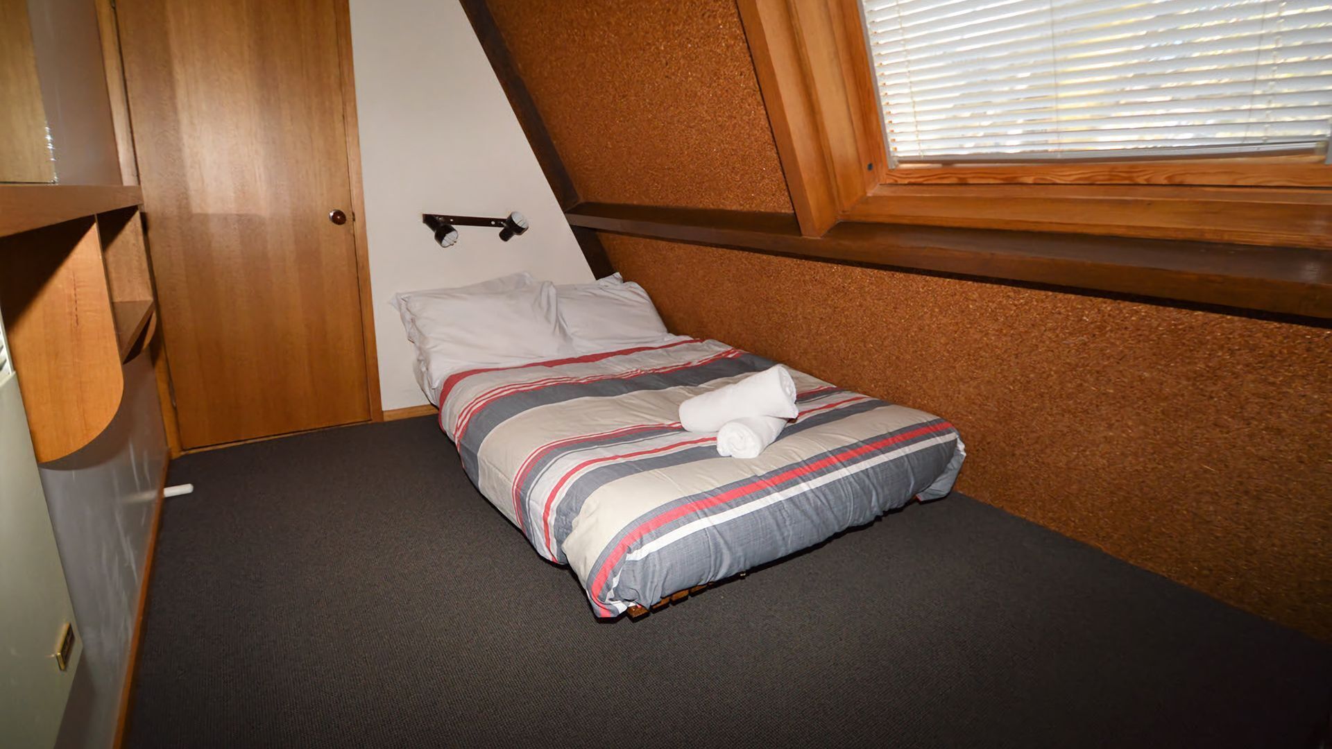 A single bed with a striped bedspread and a rolled towel, a wooden headboard, a small shelf, and a window with blinds.