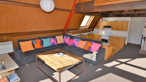 A cozy living area with a sectional sofa, a wooden coffee table, and a kitchenette in the background.