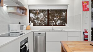 A modern kitchen with white cabinetry, a stainless steel dishwasher, a microwave, and a window with a view of trees.