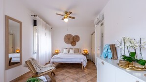 Un dormitorio con cama, lámparas de noche, ventilador de techo, espejo, silla, mesita de noche con cesta y cómoda con objetos decorativos.