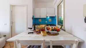 Una cocina pequeña y luminosa, con armarios blancos, salpicadero de azulejos azules, y una mesa de comedor dispuesta con comida y vino.