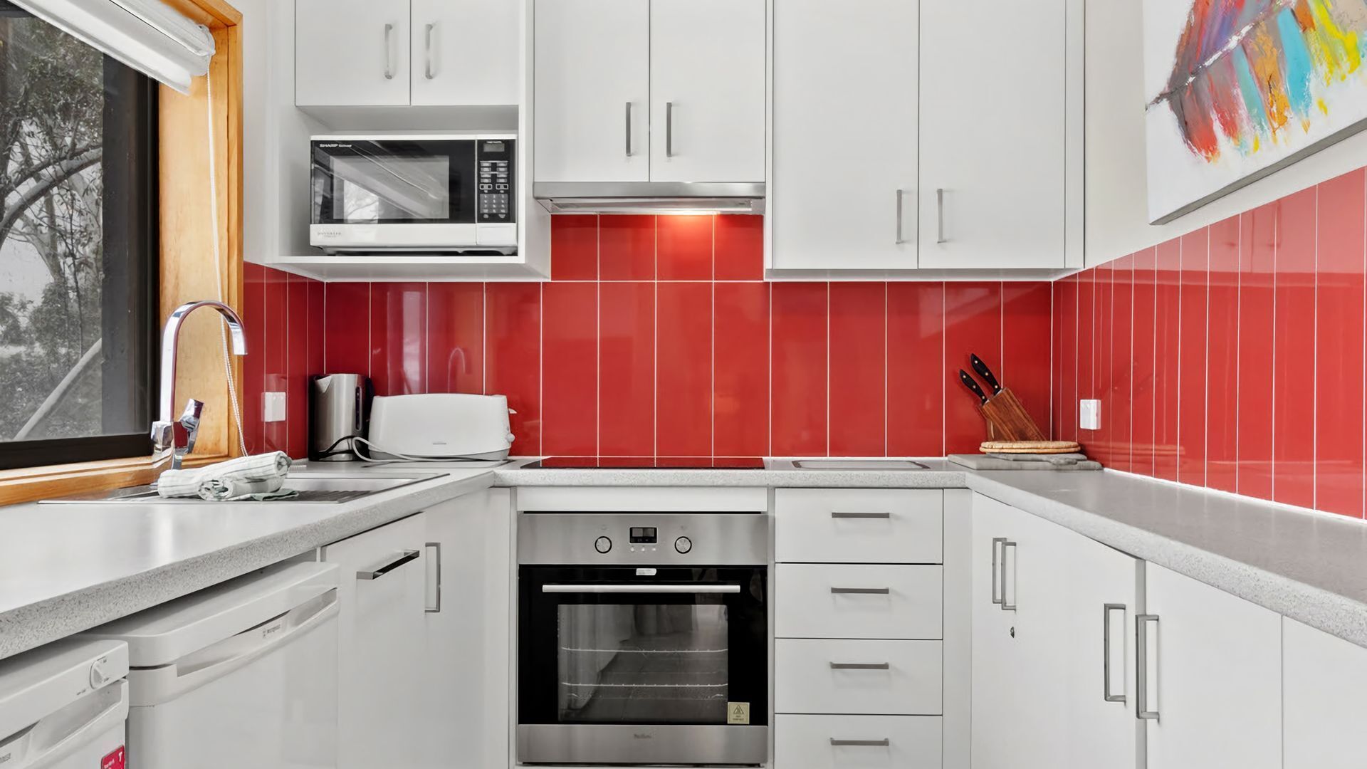 Una cucina moderna con mobili bianchi, un forno a microonde, un tostapane, un lavello e uno schienale piastrellato rosso.