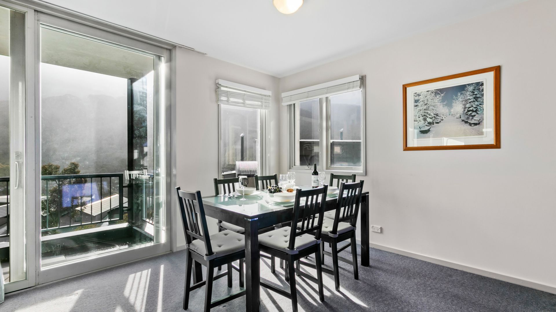 Un coin repas avec une table et des chaises en verre, de grandes fenêtres et un tableau encadré au mur.
