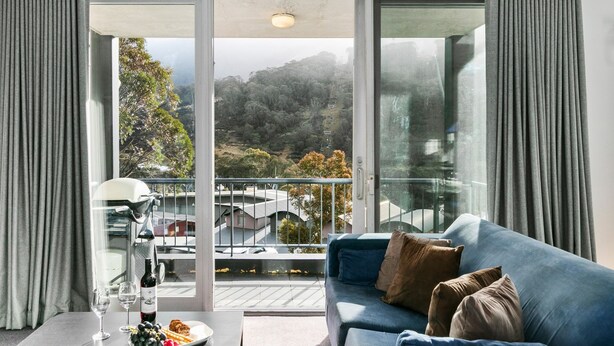 Un salon moderne avec un canapé bleu, une table à manger agrémentée de verres à vin et d’une bouteille, et un balcon offrant une vue sur les arbres et les bâtiments.