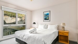 Une chambre avec un grand lit, deux tables de chevet, une fenêtre avec des stores et un tableau encadré au mur.