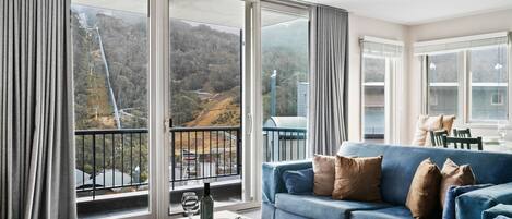 A living room with a blue sofa, a glass coffee table, and a view of a mountainous landscape through large windows.