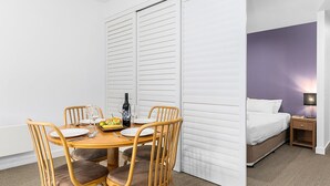 A hotel room with a dining area featuring a wooden table and chairs, a bed with white bedding, and a purple accent wall.
