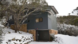 Una casa moderna de dos pisos con base de piedra y revestimiento oscuro, rodeada de árboles y colinas cubiertas de nieve.