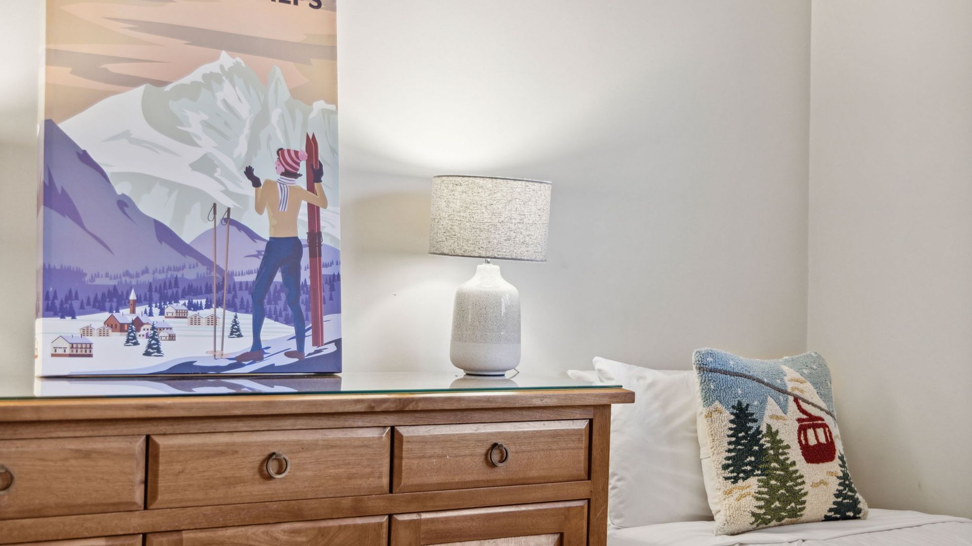 Une chambre à coucher avec une commode en bois, une lampe, un oreiller et un tableau représentant un paysage de montagne enneigé.