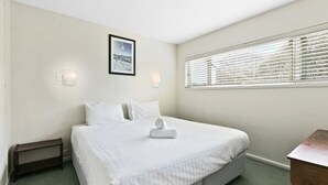 Una habitación de hotel con una cama, una mesita de noche de madera, una ventana con persianas y un cuadro en la pared.