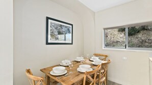 Comedor con mesa de madera dispuesta para cuatro, platos blancos y copas de vino. Cuatro sillas de madera rodean la mesa. Un cuadro enmarcado cuelga de la pared sobre la mesa.