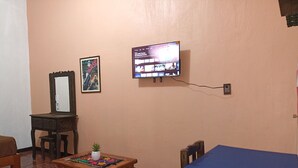 Una habitación con un televisor montado en la pared, una mesa de madera con un tapete colorido, una pequeña planta en maceta y un mantel azul sobre una mesa.