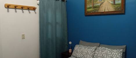 A bedroom with a bed, a blue curtain, a wooden coat rack, a painting on the wall, and a bedside table with a lamp.
