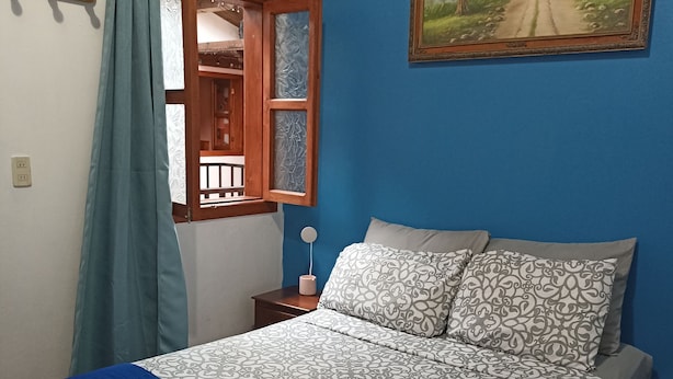 Un dormitorio con cama, mesita de noche, ventana con cortinas, un cuadro en la pared y una manta azul sobre la cama.
