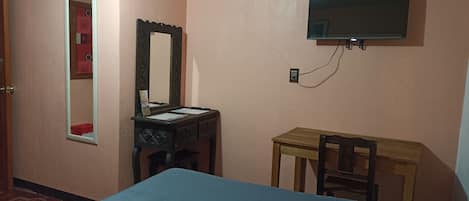 A bedroom with a bed, a desk, a chair, a mirror, and a television mounted on the wall.