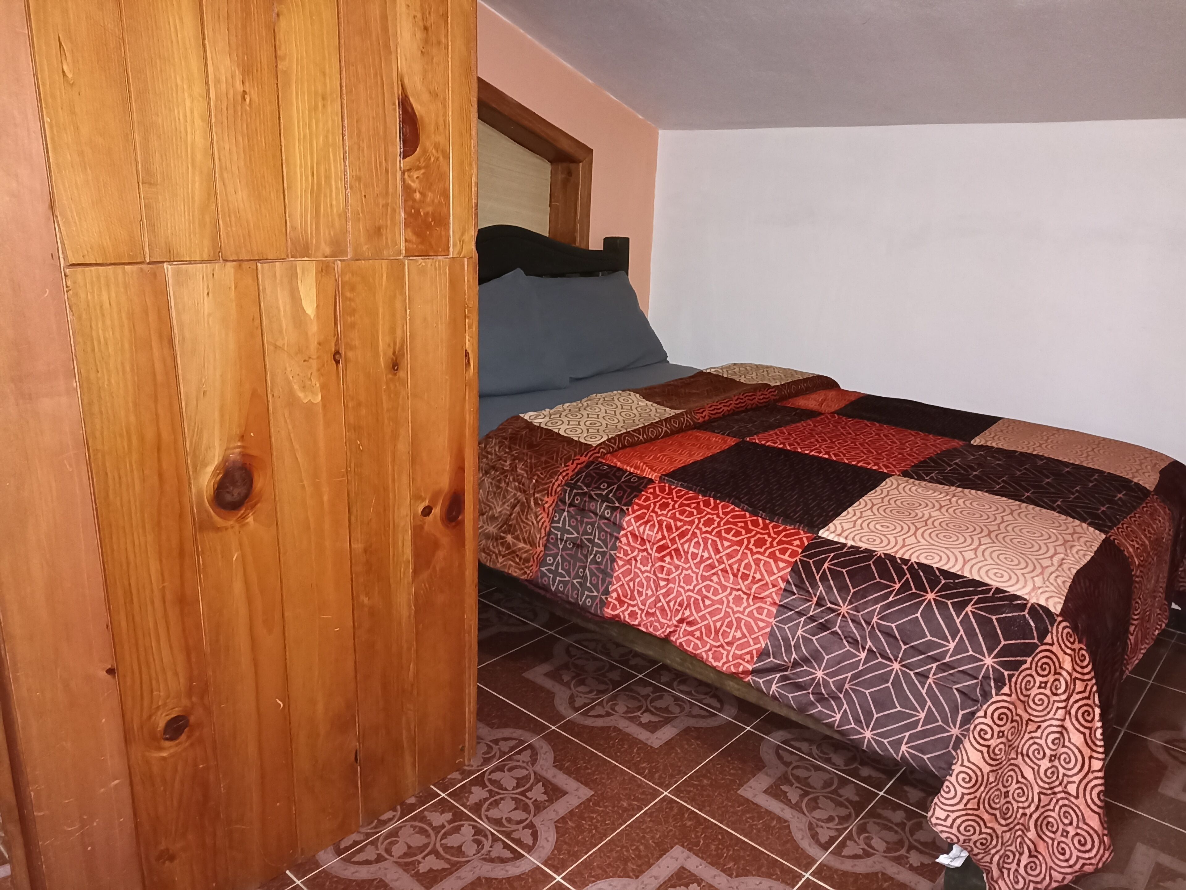 A bedroom with a wooden wardrobe, a patterned bedspread, and a tiled floor.
