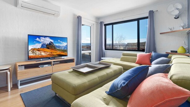 A modern living room with a flat-screen TV, a sofa set, and a large window offering a view of the outdoors. - Aspen Court 8 is a comfortable and spacious apartment (Jindabyne)