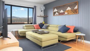 A modern living room with a sectional sofa, a small table, and a view of the outdoors through a large window. - Aspen Court 8 is a comfortable and spacious apartment (Jindabyne)