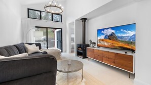 Ein modernes Wohnzimmer mit einem großen Flachbildfernseher, einem schwarzen Kaminofen, einem grauen Sofa und Blick auf Berge durch das Fenster.
