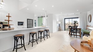 Eine moderne Küche mit weißer Arbeitsplatte, einer eingebauten Mikrowelle und einem Kühlschrank mit zwei Türen. Drei schwarze Hocker mit gewebten Sitzflächen und ein Holztisch mit Stühlen sind ebenfalls vorhanden.