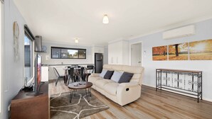 Una sala de estar moderna con un sofá color crema, una mesa de centro y un comedor con sillas, con una cocina al fondo.