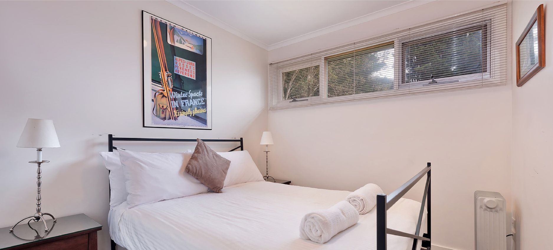Una cama bien hecha con sábanas blancas, una sola almohada marrón y una toalla blanca enrollada al lado. Hay un cartel en la pared sobre la cama, una mesita de noche con una lámpara y una ventana con persianas.