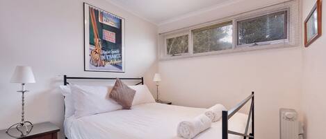 Una cama bien hecha con sábanas blancas, una sola almohada marrón y una toalla blanca enrollada al lado. Hay un cartel en la pared sobre la cama, una mesita de noche con una lámpara y una ventana con persianas.