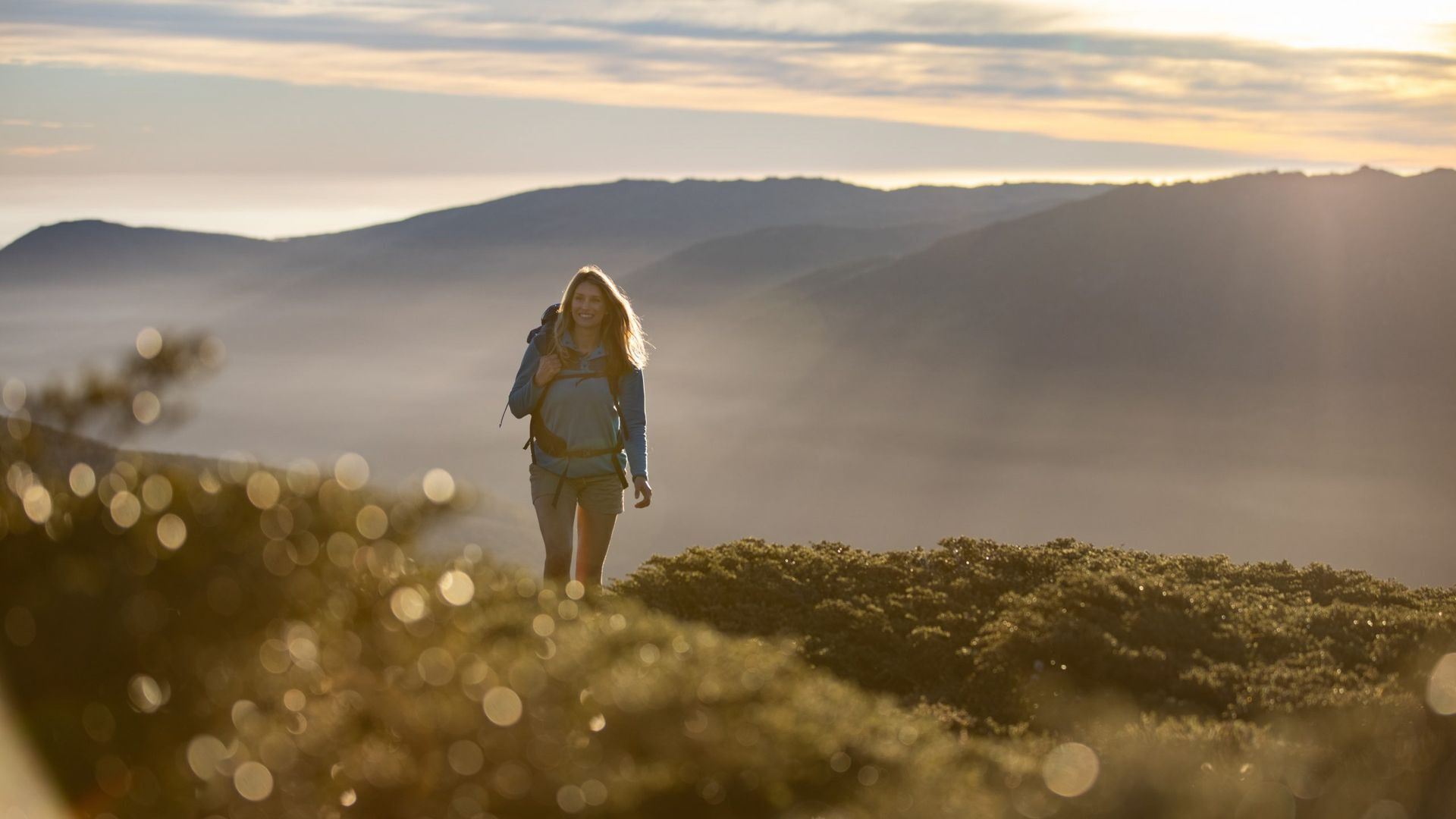Una persona caminando por un sendero de montaña, con una vista despejada de montañas lejanas y un amanecer o atardecer al fondo.