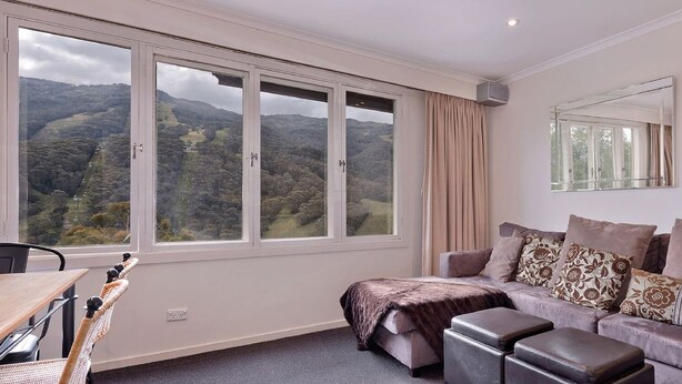 A room with a large window, a sofa, a table, and a mirror. - Karoonda 6 is warm and welcoming three bedroom apartment. (Thredbo)