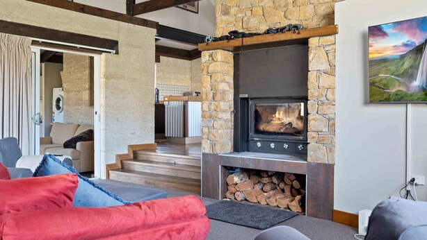 A cozy living room with a stone fireplace, a staircase leading to another level, and a painting of a waterfall on the wall. - Old Hut Creek Chalet with a peaceful rural outlook (Jindabyne)