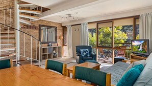 Ein modernes Wohnzimmer mit einer Holztreppe, einem großen Fenster mit Blick auf Bäume, einem Fernseher und einem Essbereich mit Tisch und Stühlen.