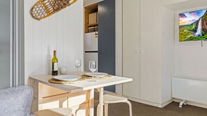 Quarto compacto com uma mesa de jantar posta para dois, uma garrafa de vinho e duas taças. Uma televisão está fixada na parede e uma cadeira é visível em primeiro plano.