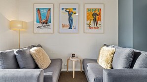 Uma sala de estar moderna com dois sofás cinzentos, uma pequena mesa de apoio de madeira e três cartazes emoldurados na parede.